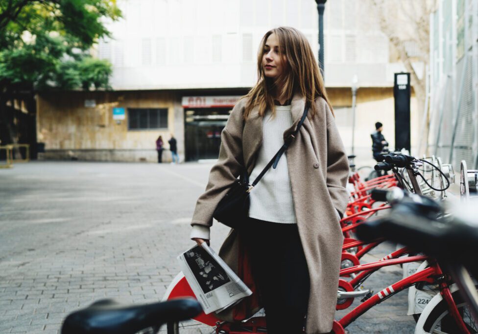 Charming hipster girl choosing the city bike for the rent on a blurred street background. Attractive caucasian woman looking aside while standing with a newspaper in her hand amidst the city bicycles.